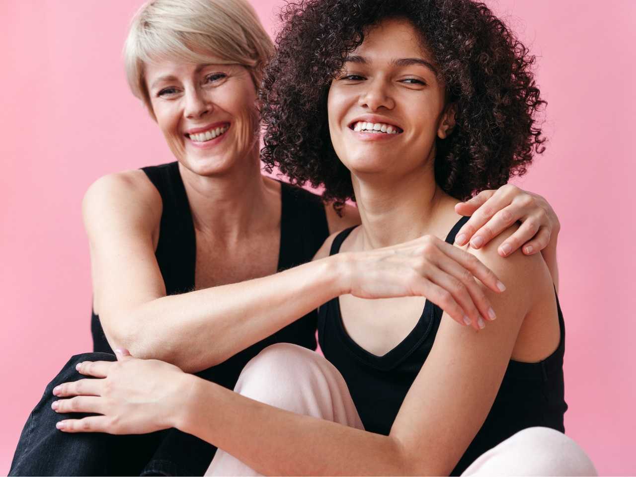 Two women hugging against pink background.